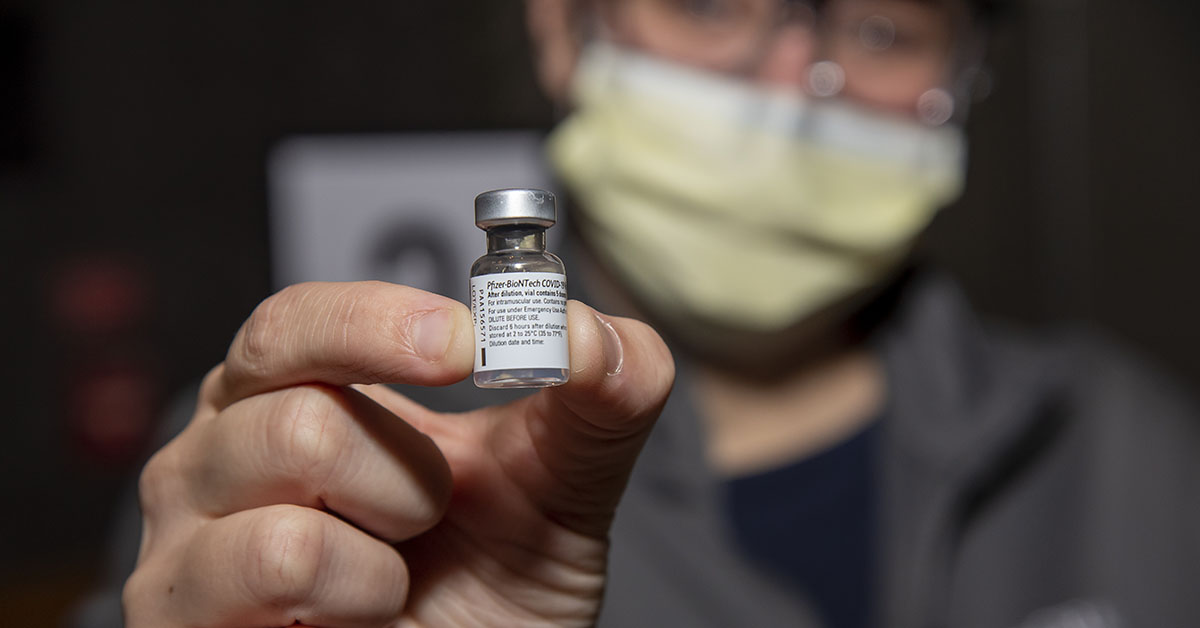 A photograph with a blurred background of a person holding a vial that reads "Pfizer-BioNTech COVID-19 Vaccine".