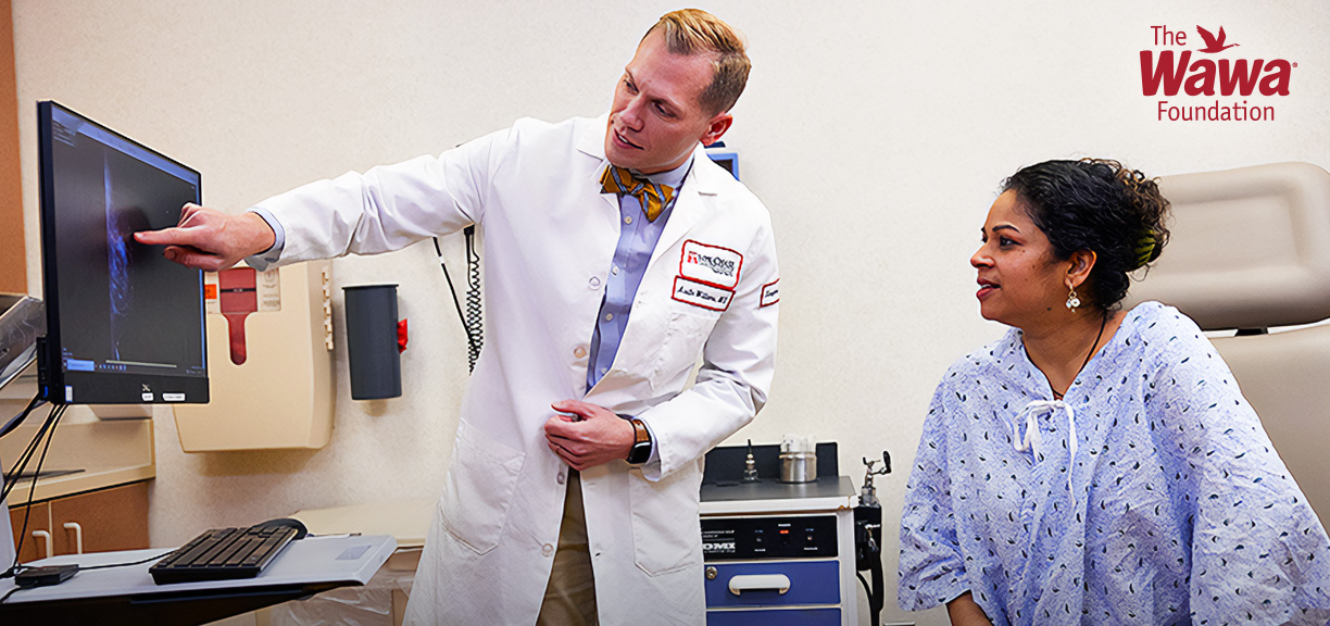 A healthcare provider in a white lab coat points to a medical image displayed on a computer monitor while discussing the results with a patient seated in an exam room