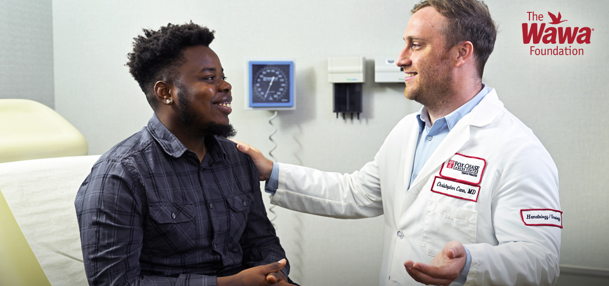 A doctor in a white lab coat sits beside a patient in an exam room, smiling and speaking with him while placing a reassuring hand on his shoulder