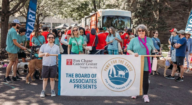 Participants and their dogs gather at an outdoor community event while two people hold a banner reading “Fox Chase Cancer Center Temple Health – The Board of Associates Presents Paws for the Cause.”