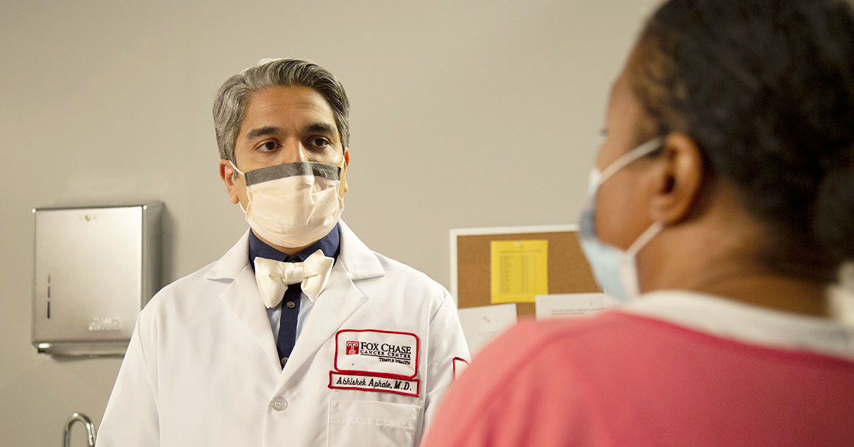 A photograph looking over a patient's shoulder as they talk to a Fox Chase doctor in a medical room, both wearing masks.