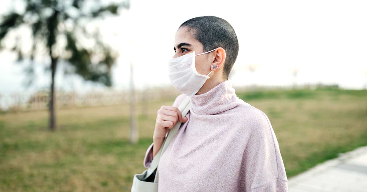 A photograph of a feminine person wearing a mask, standing outside and looking off to the side.