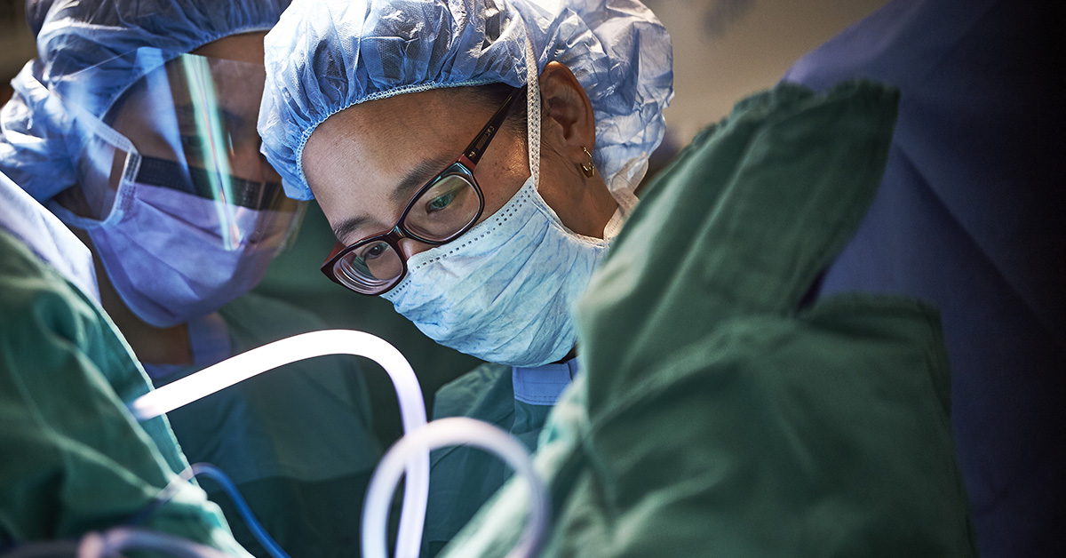 A closeup photograph of several medical professionals in full surgical garb, surrounding the same thing as they work.