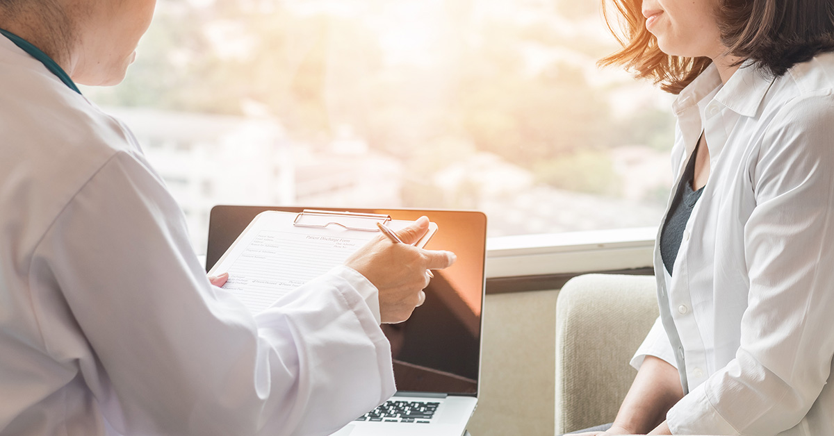 A photo from behind two people, with a clipboard and laptop in front of them and bright sunlight coming in from the window.