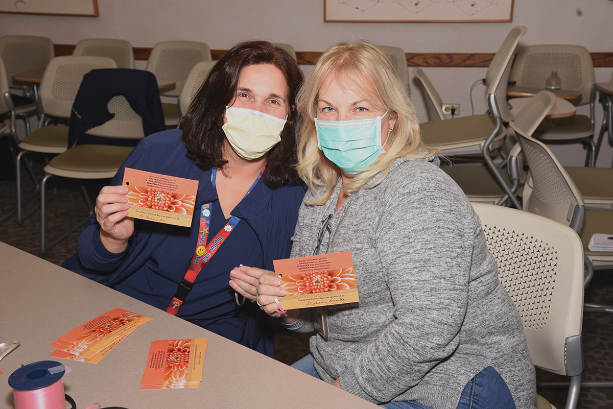 Two women in masks holding orange cards with encouragement and a name of the sponsor of the Chemo Comfort Bag