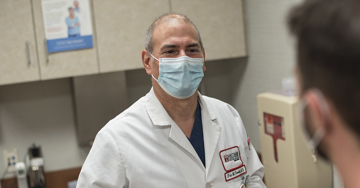 A photo looking over the shoulder of a patient as a masked Fox Chase doctor looks at them, both standing in a medical room.