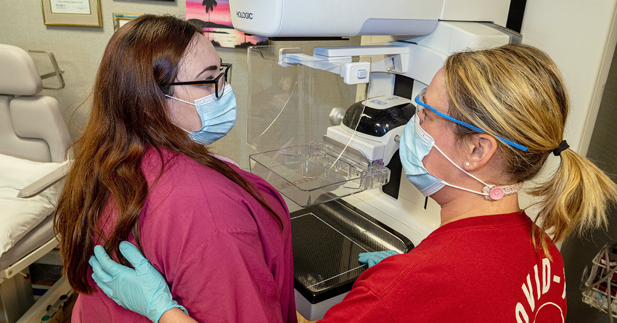 A medical professional and a patient stand in front of a mammogram machine, wearing masks and looking at each other.