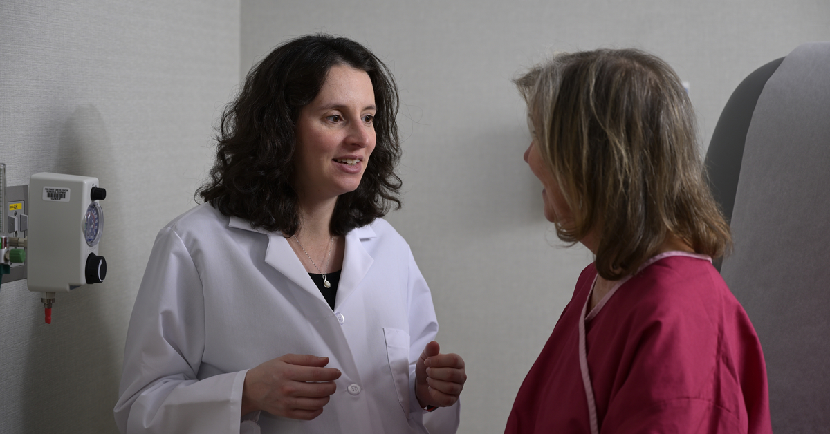 A photograph of a medical professional standing close to a patient in a medical chair, as they smile and speak with each other.