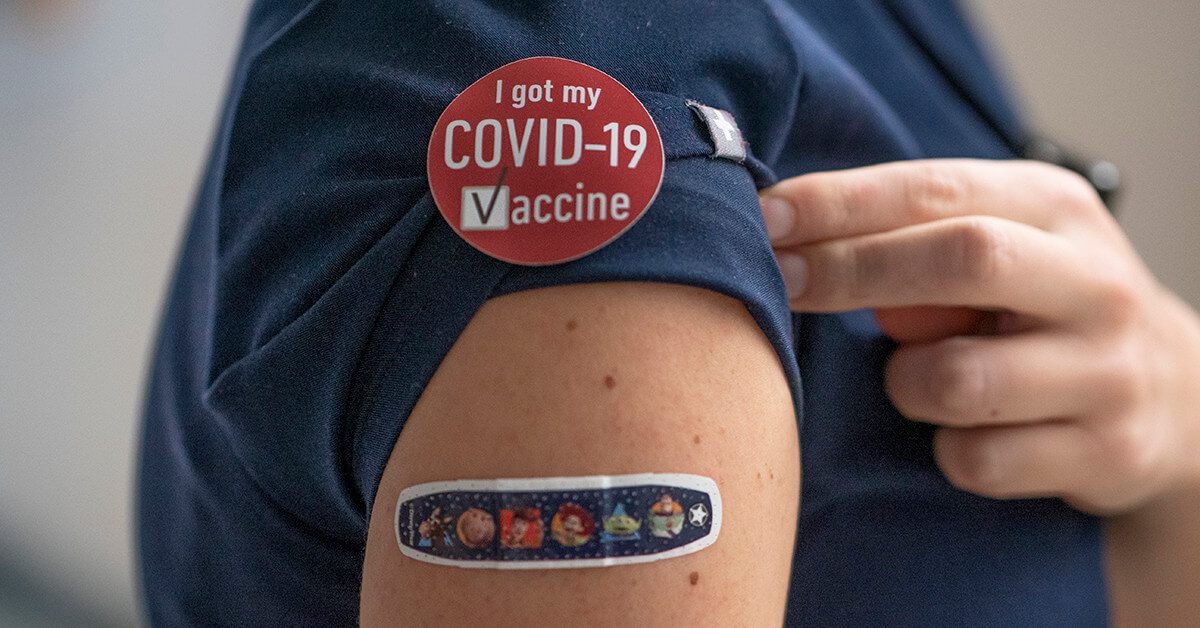 A close-up photograph of a bandaid on a person's upper arm, with a sticker that says "I got my COVID-19 vaccine" on their shirt.