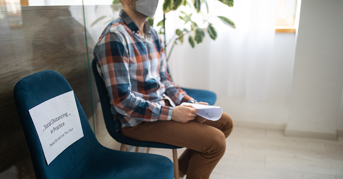 A photograph of a person sitting in a teal chair in the corner of a room, holding a paper and looking off camera.