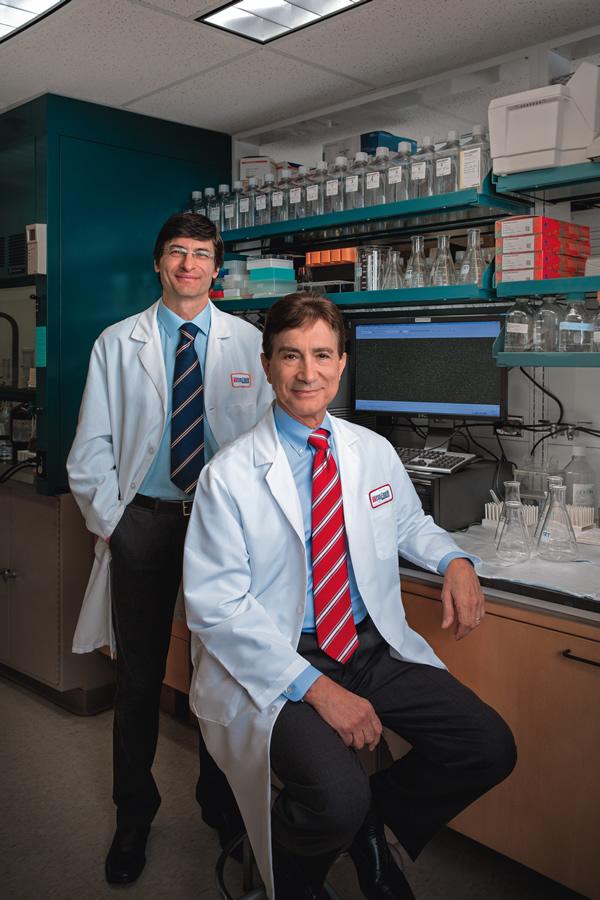 A photo of Alfonso Bellacosa (left, standing up) and Joseph R. Testa (right, sitting on a stool), in a laboratory.