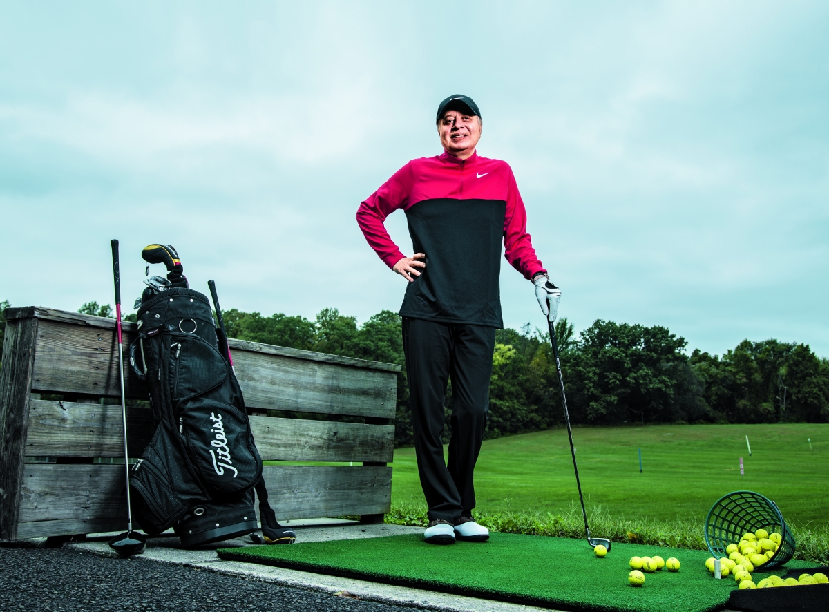 A photo of Mahmood Saeed standing on a golf course with an iron in his hand, with his clubs next to him and several balls on the ground.