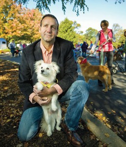 A photograph of Frank Serianni outside with several other people in the background, kneeling down and holding his white dog.