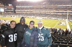 A photo of four people smiling at the camera, with a football game in a large stadium in the background.