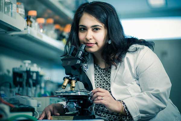 A photograph of Dr. Sanjeevani Arora, PhD sitting in a laboratory and holding a microscope close to her.