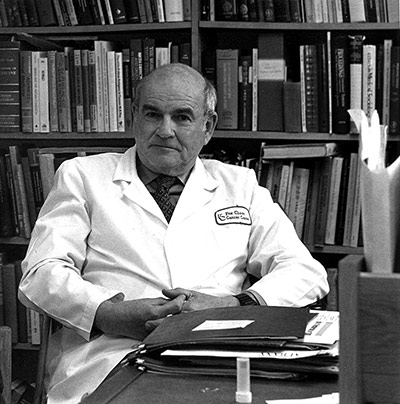 A black and white photograph of Nobel Laureate Baruch S. Blumberg, who discovered the Hepatitis B virus, sitting in a room full of books.