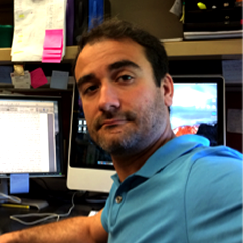 A closeup photo of Alexander Deneka, MD, PhD, turning around to look at the camera with several monitors on a desk behind him.