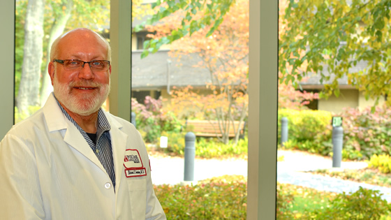 A photograph of Dr. Richard E. Greenberg standing in front of a tall glass window, smiling at the camera.