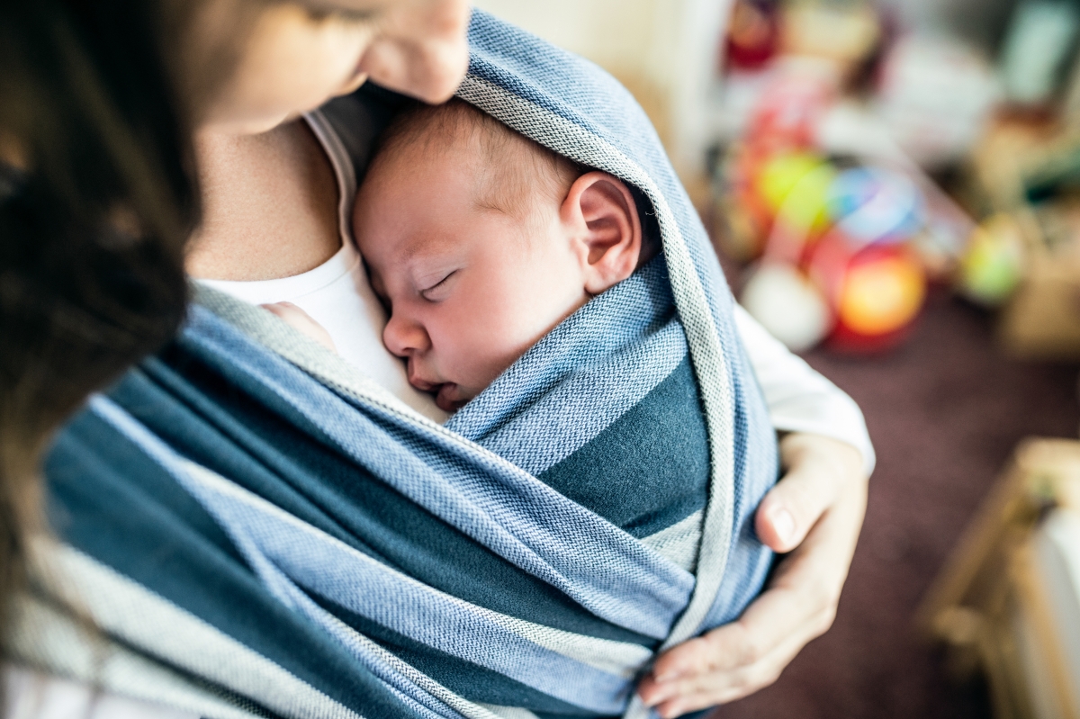 A closeup photo of a baby nestled against a woman's chest, held close by a blanket wrapped around both of them.