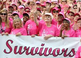 A photograph of a crowd of women wearing pink shirts, holding a sign in front of them that says "Survivors" in pink cursive.