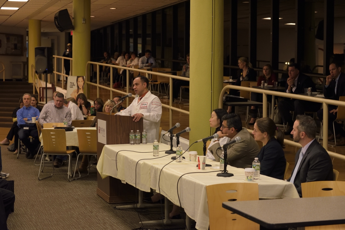 A side view photograph of a Fox Chase doctor speaking to a crowd at a podium, with people at tables on either side of him.