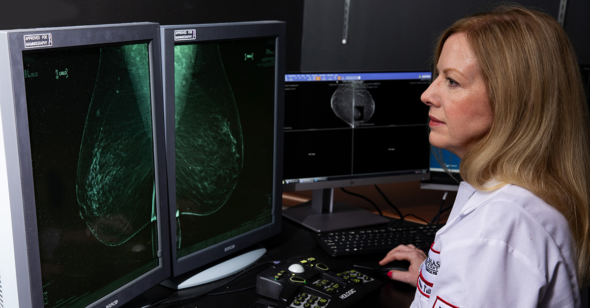 A photograph of a medical professional looking at two screens with images from a mammogram displayed on both.