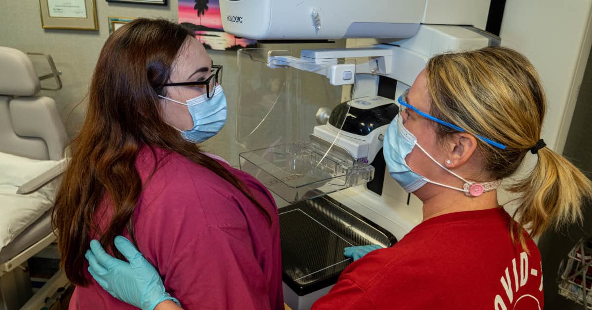 A medical professional and a patient stand in front of a mammogram machine, wearing masks and looking at each other.