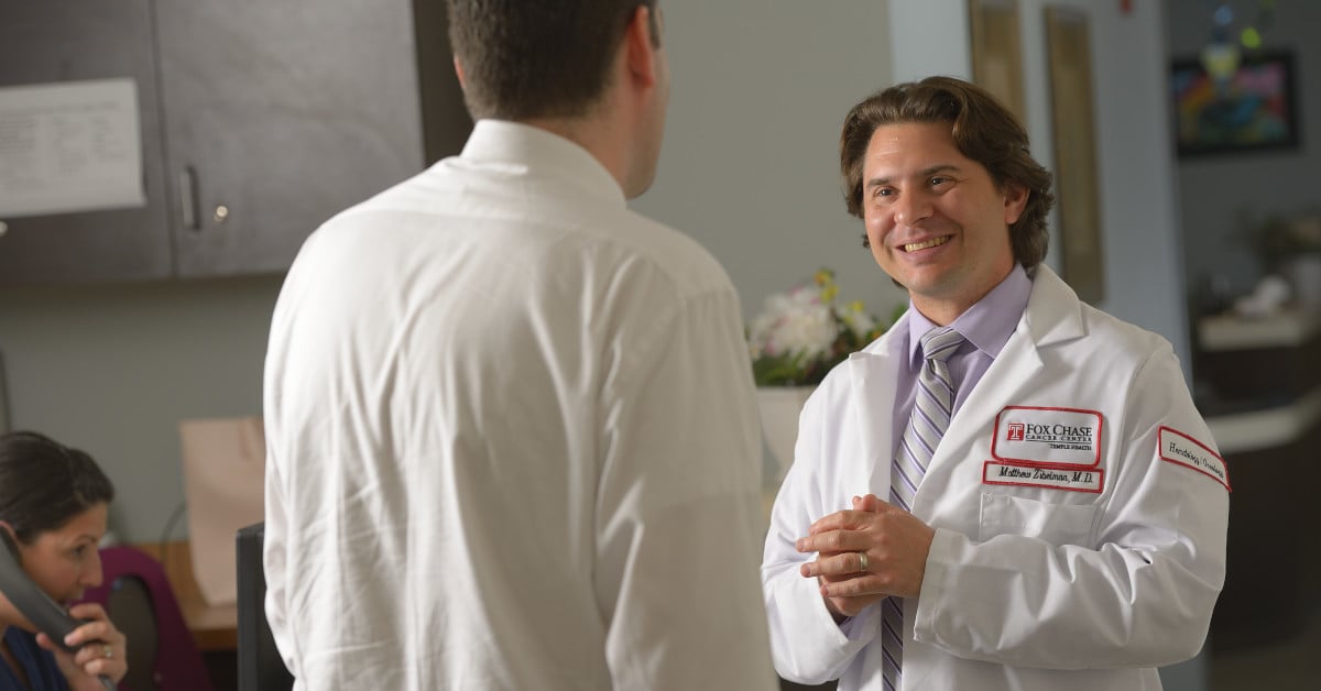 A photograph of a Fox Chase doctor smiling widely at someone, with another person on the phone at a desk to their left.