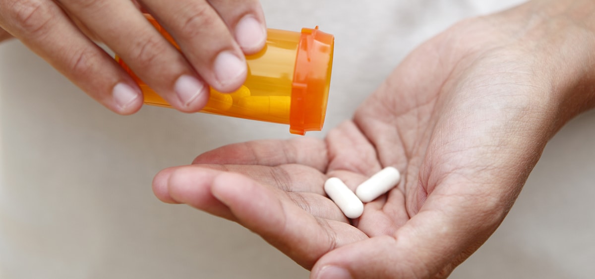 A closeup photo of two white capsules being shaken from a pill bottle in one hand into the other.