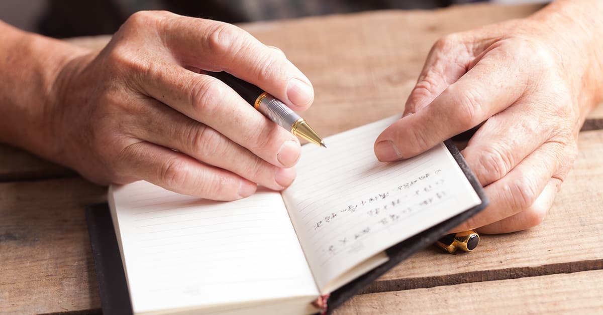 A closeup photo of a person's hands, holding a pen as they write in a small notebook.