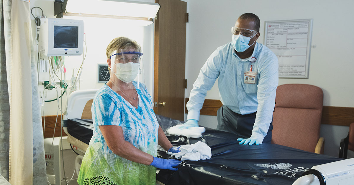 A photograph of two people sanitizing a patient room, one looking up and smiling at the camera.