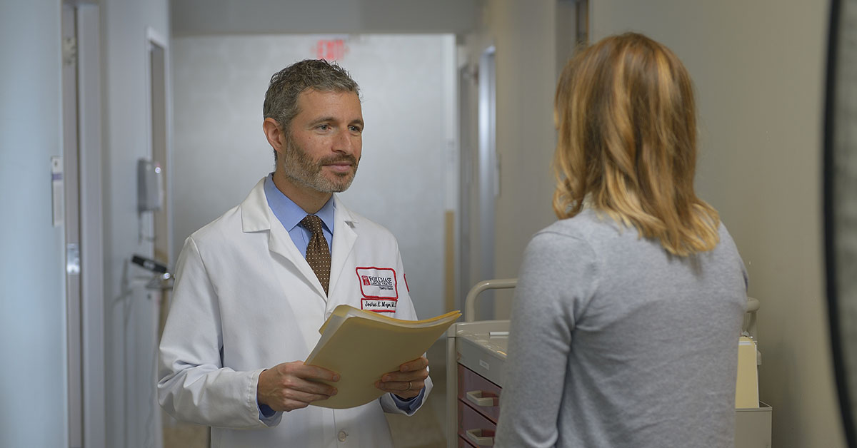 A Fox Chase doctor stands facing another person in a hallway, holding a file in his hands.