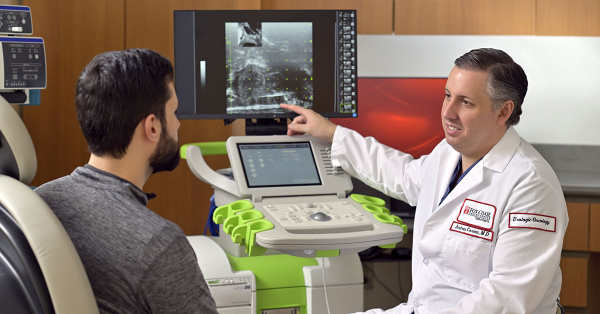 A photograph of a Fox Chase doctor speaking to a patient in a medical chair while gesturing at an image on a screen.