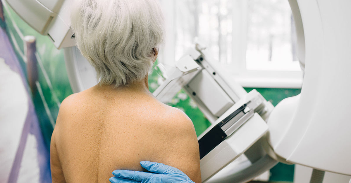 A photograph from behind a person receiving a mammogram, with a gloved hand on their back.