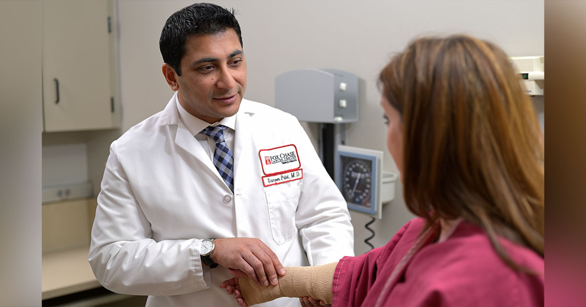 A Fox Chase doctor smiles as he examines a patients arm, which is covered in a tan medical wrap.