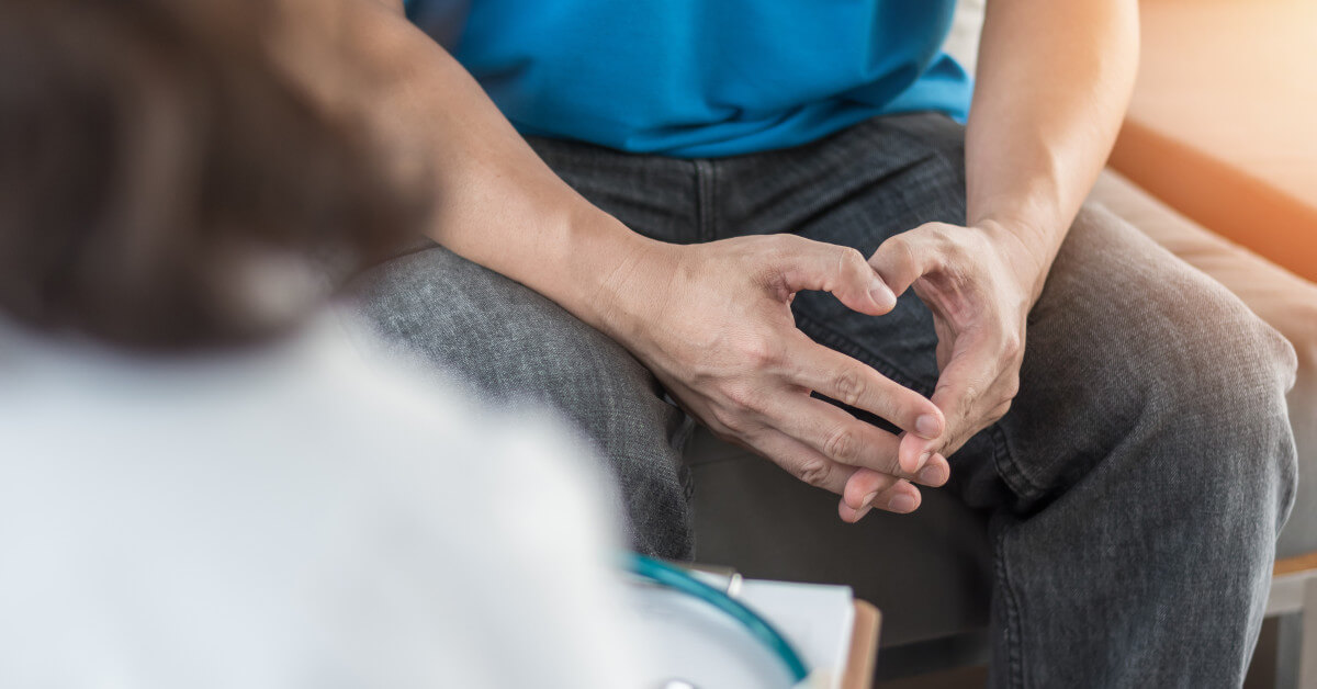 A closeup photo looking over a doctor's shoulder to a person's lap, where they loosely intertwine their fingers together.