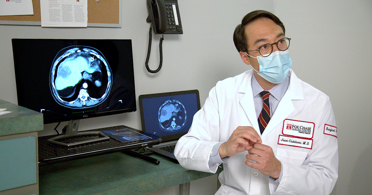 A photograph of a Fox Chase doctor looking off camera, with two screens on a desk to his left showing medical imaging results.