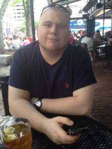 A photograph of  Andrew Steinmetz sitting at an outdoor table, with a phone in hand and a drink in front of him.