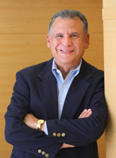 A half body photo of a well dressed man, smiling at the camera as he leans against the wall.
