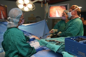 A photograph of three doctors performing surgery, looking up at two screens showing the feed from a laparoscope.