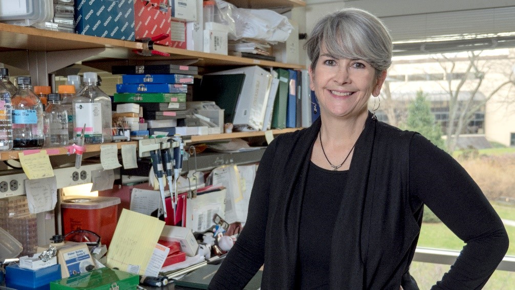 A photograph of Denise Connolly standing in a cluttered laboratory, smiling at the camera.