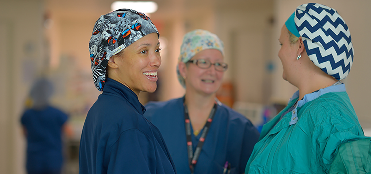 A photograph of three medical professionals wearing hair coverings smiling as they speak to each other in a hospital hallway.