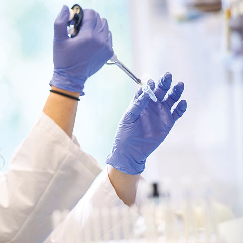 A close up photo of two gloved hands using a long cylindrical medical tool to put liquid into a small vial.