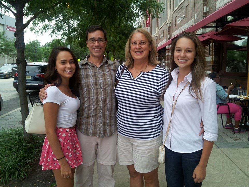 A photograph of a family of four smiling at the camera on a street lined with shops, a mother, father, and two teenage daughters