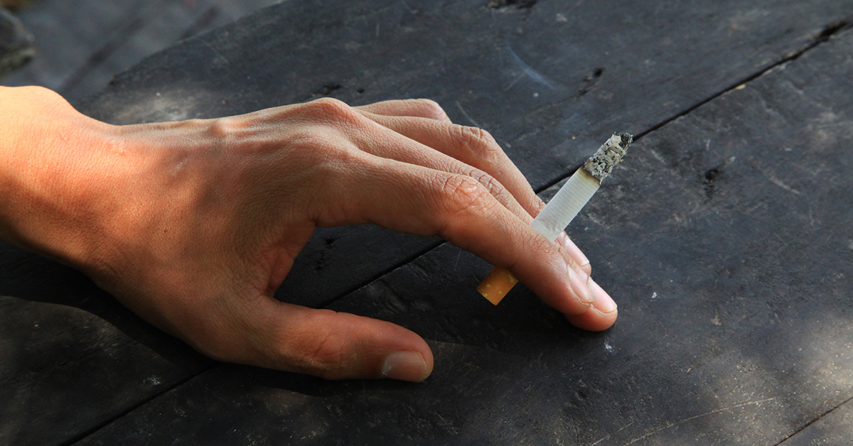 A closeup photo of a person's hand resting on a dark stone surface, holding a cigarette between their fingers.