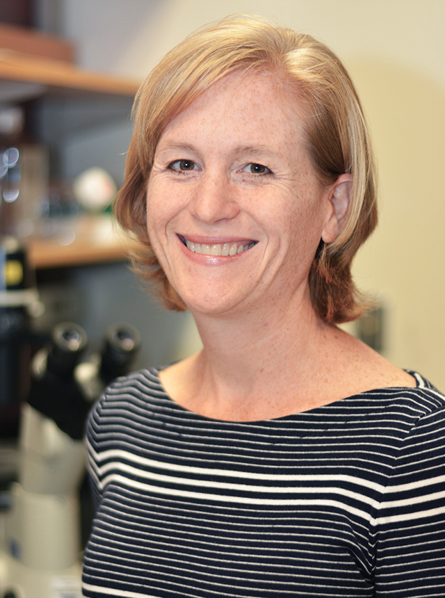 A portrait shot of Tiffiney Hartman standing in a laboratory, smiling at the camera.