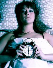 A photograph of a person laying in bed in a dark room, looking up at the ceiling with an alarm clock in their hands.
