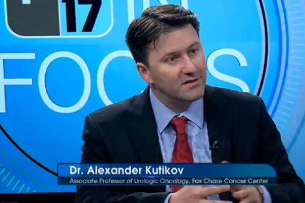 Dr. Alexander Kutikov on a TV show with the channel's logo in the background, speaking to someone off camera.