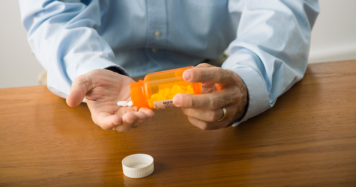 A photo of a person leaning over a wooden surface as they shake three tablets from a pill bottle into their other hand.