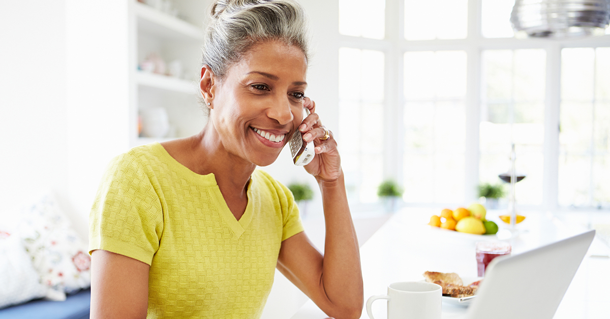 A photo of a person standing in their kitchen with a phone up to their ear and a laptop in front of them.